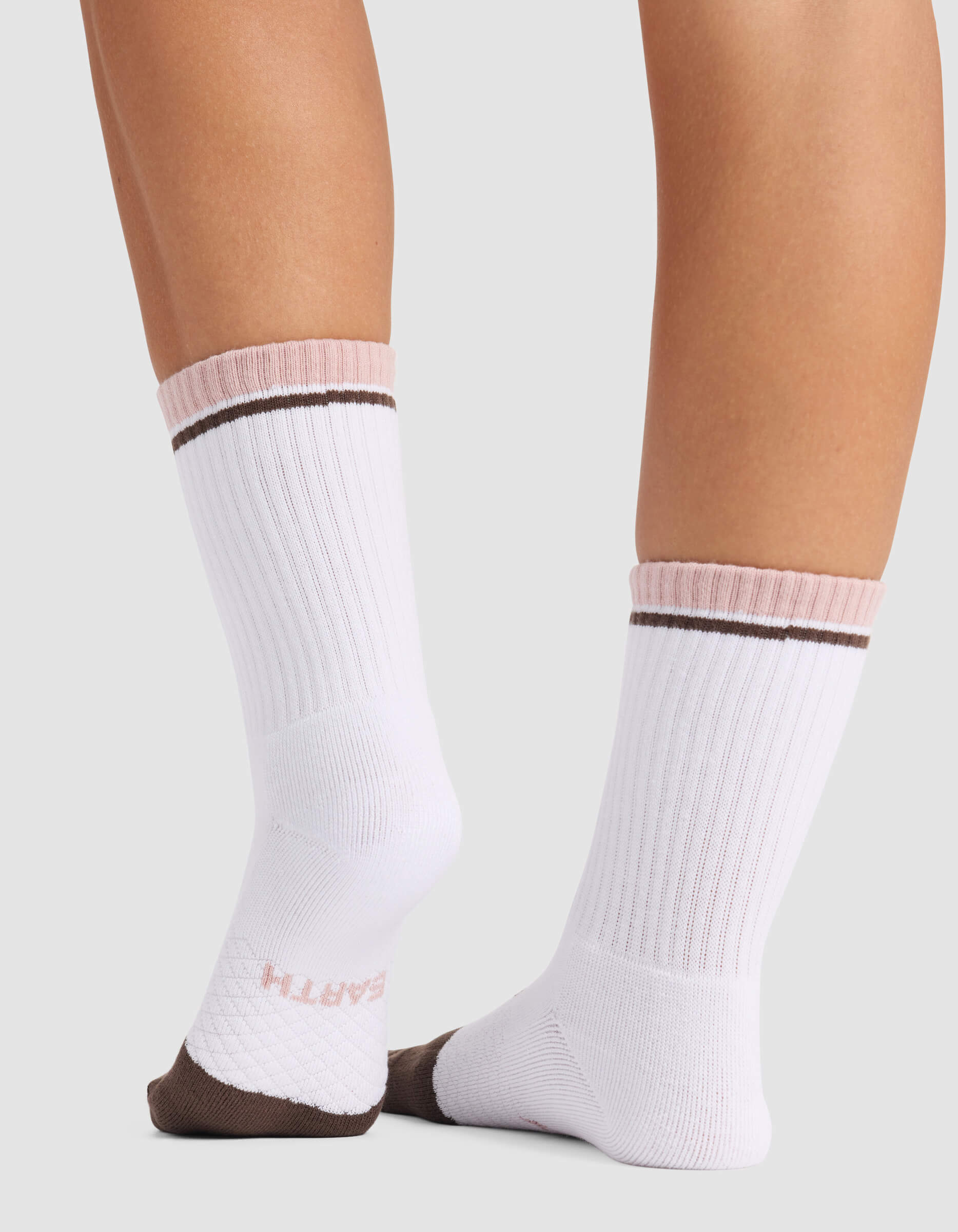 Two lower legs are shown wearing white crew socks from the Cozy Earth Women’s Essential Calf Sock 4-Pack - Last Chance, featuring pink and brown accents. The socks have a ribbed texture with “EARTH” text on the sole, set against a plain light gray background.
