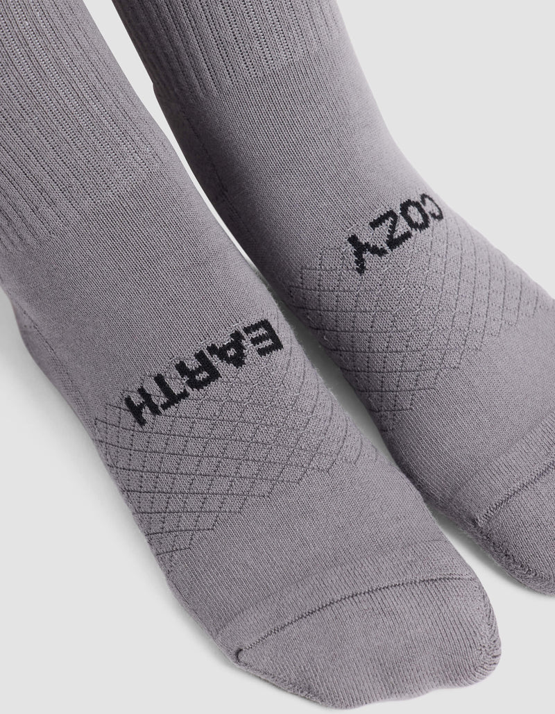 The Women's Essential Calf Sock by Cozy Earth features light gray fabric with “EARTH” on the left sock and “COZY” on the right, a ribbed cuff, and a diamond knit pattern across the top.