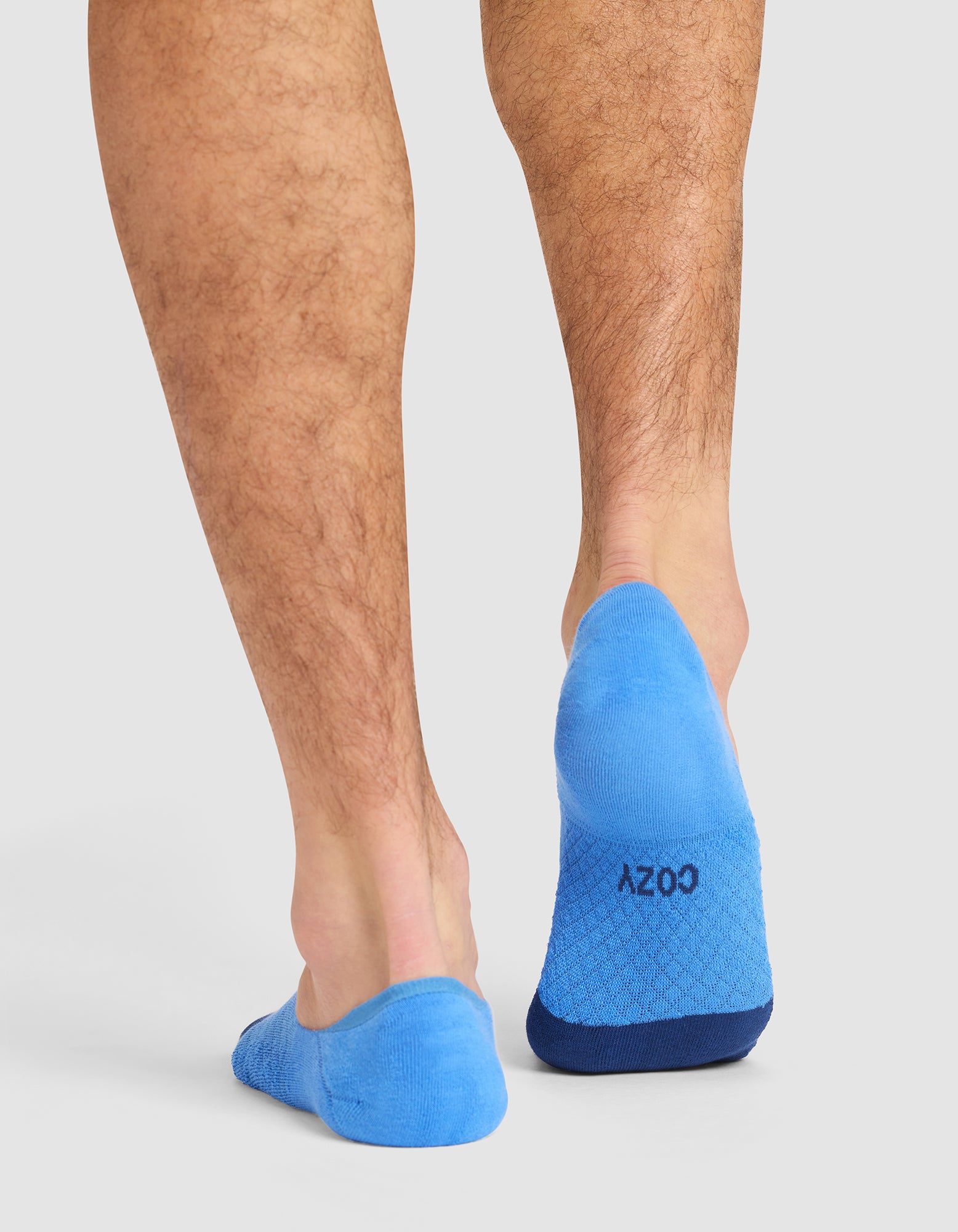 A person stands on a white background, viewed from behind, wearing bright blue Essential No-Show socks from Cozy Earth with "COZY" printed on the soles. |Color:Harbor