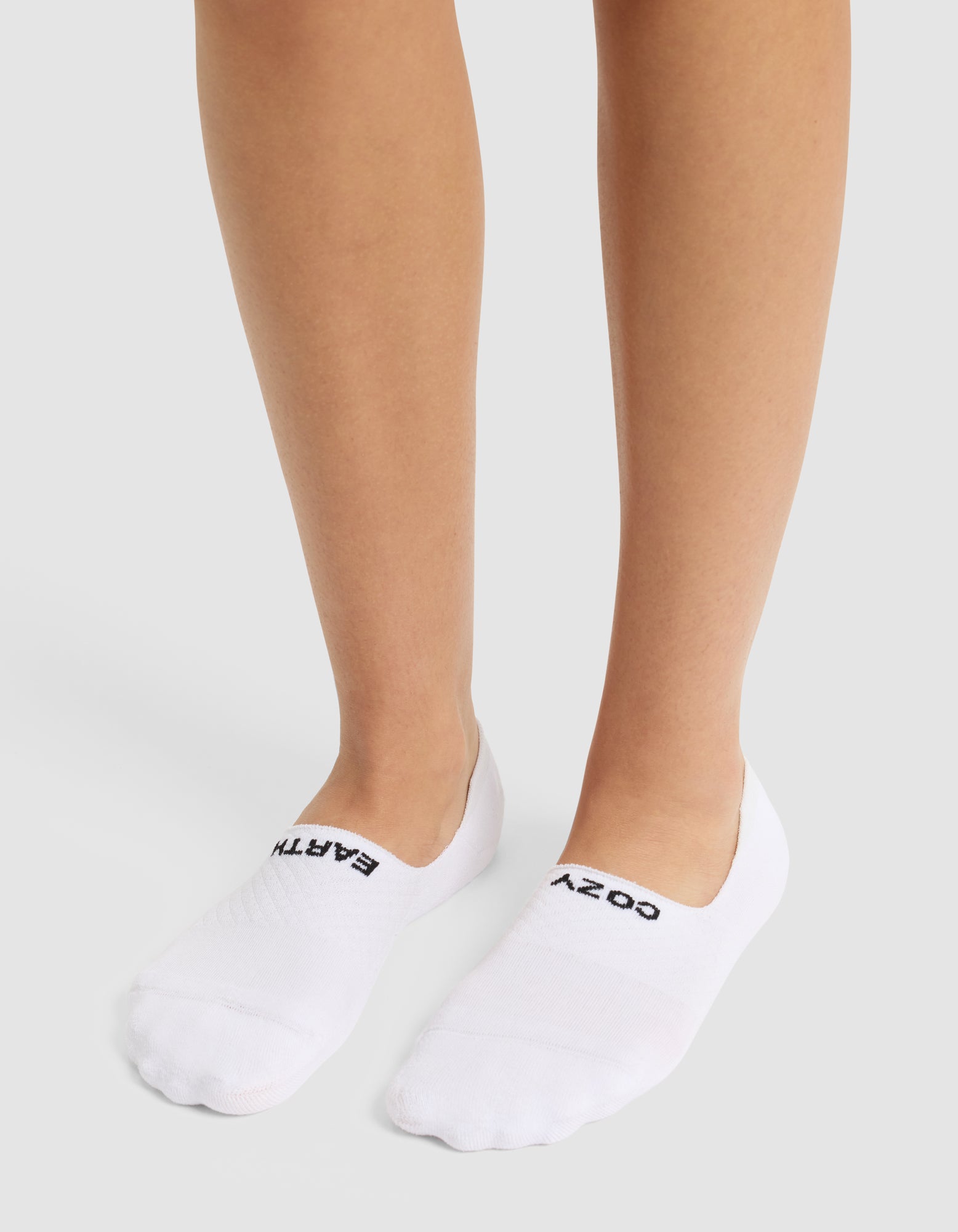 A person wears white Essential No-Show Sock 4-Pack by Cozy Earth; the left sock reads "EARTH" and the right "COZY" in black. The background is plain and light-colored.