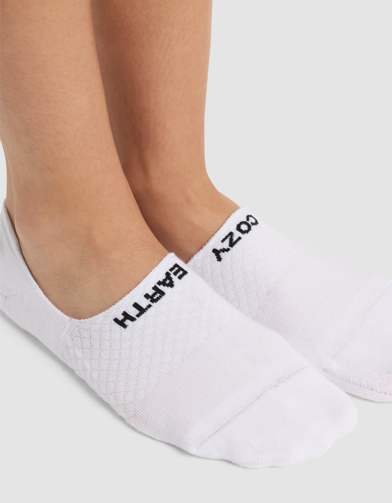 A close-up of two feet in Cozy Earth Essential No-Show Socks with black text—“EMPATH” on the left, “COZY” on the right—standing on a white surface. 