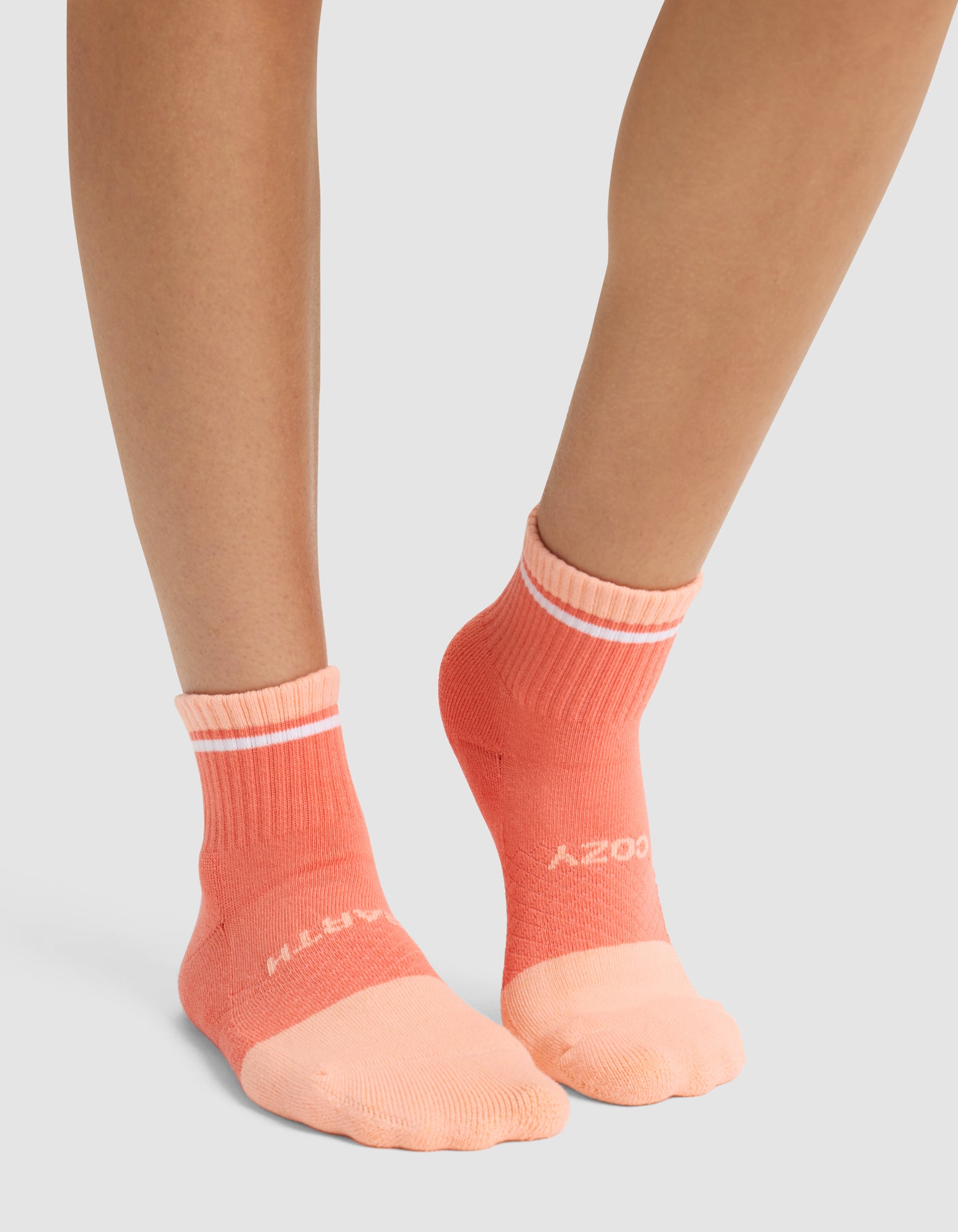 A person wearing Cozy Earth Essential Quarter Socks in peach and coral with white stripes and white front lettering, standing on a light gray surface. 