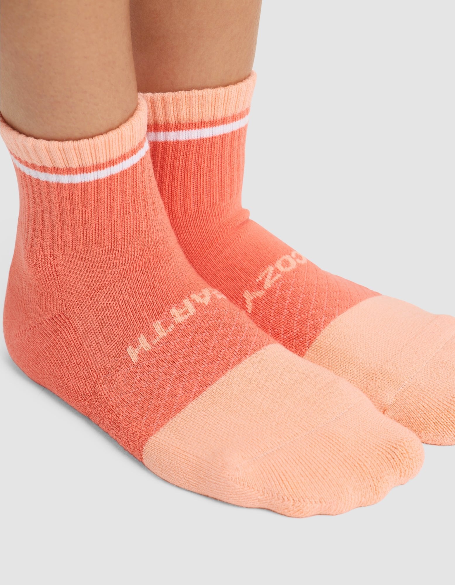 A close-up of two feet in Cozy Earth Essential Quarter Socks, coral pink with lighter pink toes, a white stripe near the cuffs, and partial text on top. Set against a light gray background. 