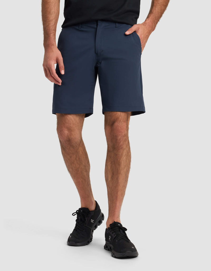 A person wearing Cozy Earth's Men's Everywhere Short in navy, a black shirt, and black athletic shoes stands with one hand in their pocket against a plain light background. Only the lower half of their body is visible.