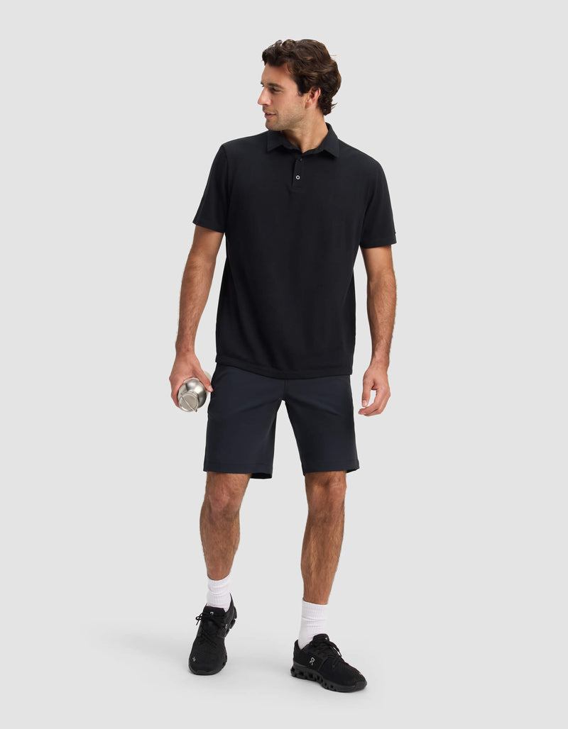 A man stands against a light gray background, wearing Cozy Earth's Men's Everywhere Short in black, paired with a black polo shirt, black sneakers, and white socks. He holds a silver water bottle and looks to his left.