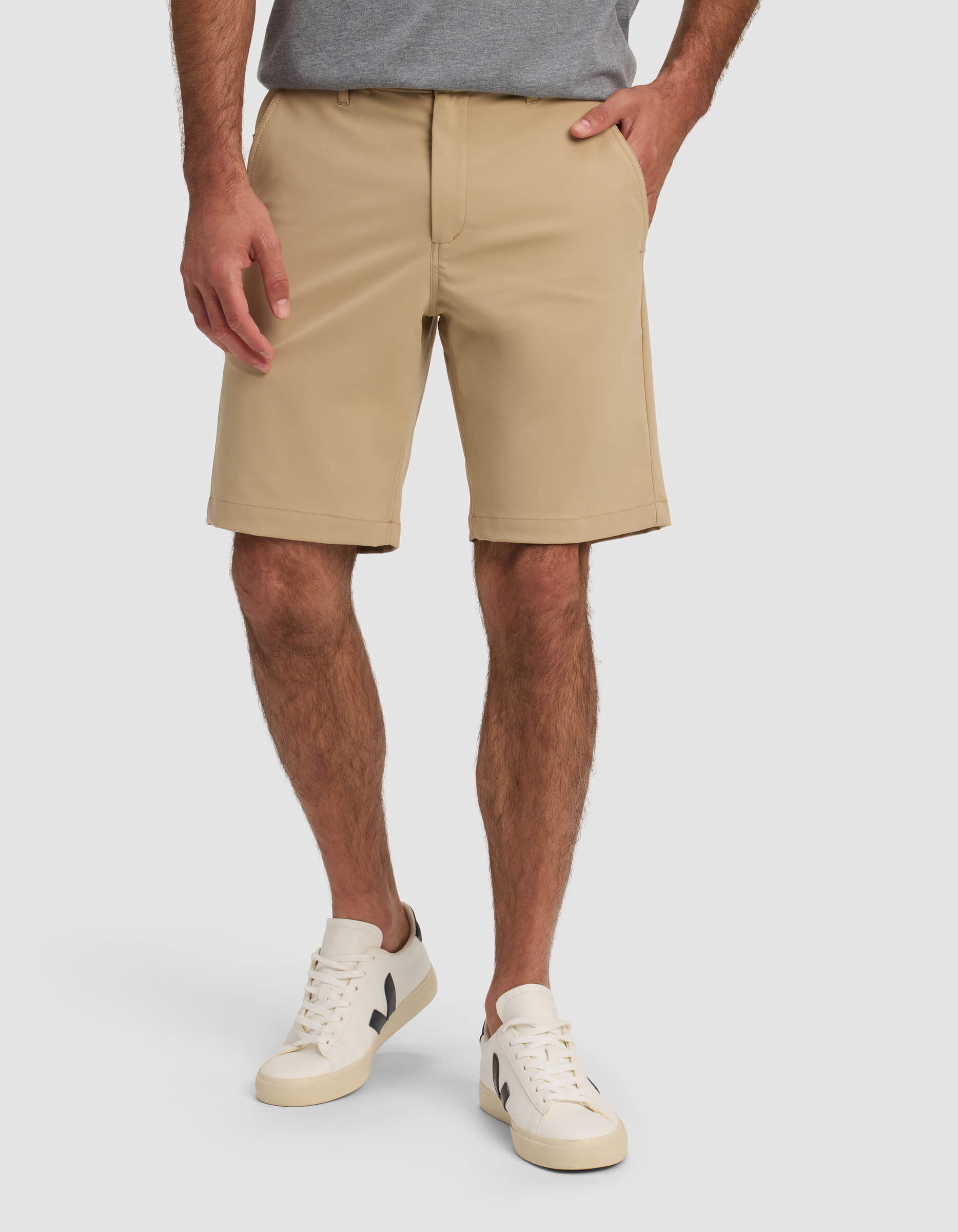 A man wearing Cozy Earth Men's Everywhere Short in beige, a gray t-shirt, and white sneakers with black accents stands with one hand in his pocket against a plain background.