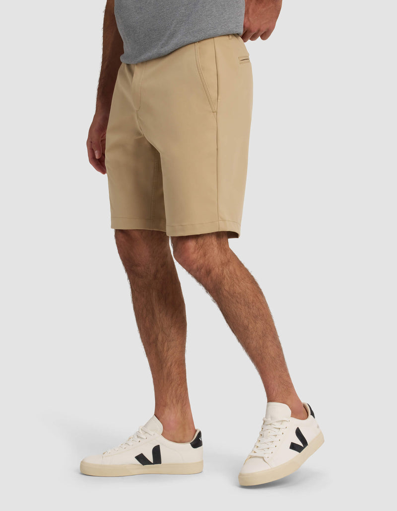 A person wears Cozy Earth's Men's Everywhere Short in beige, a gray t-shirt, and white sneakers with black detail while standing against a plain light background.