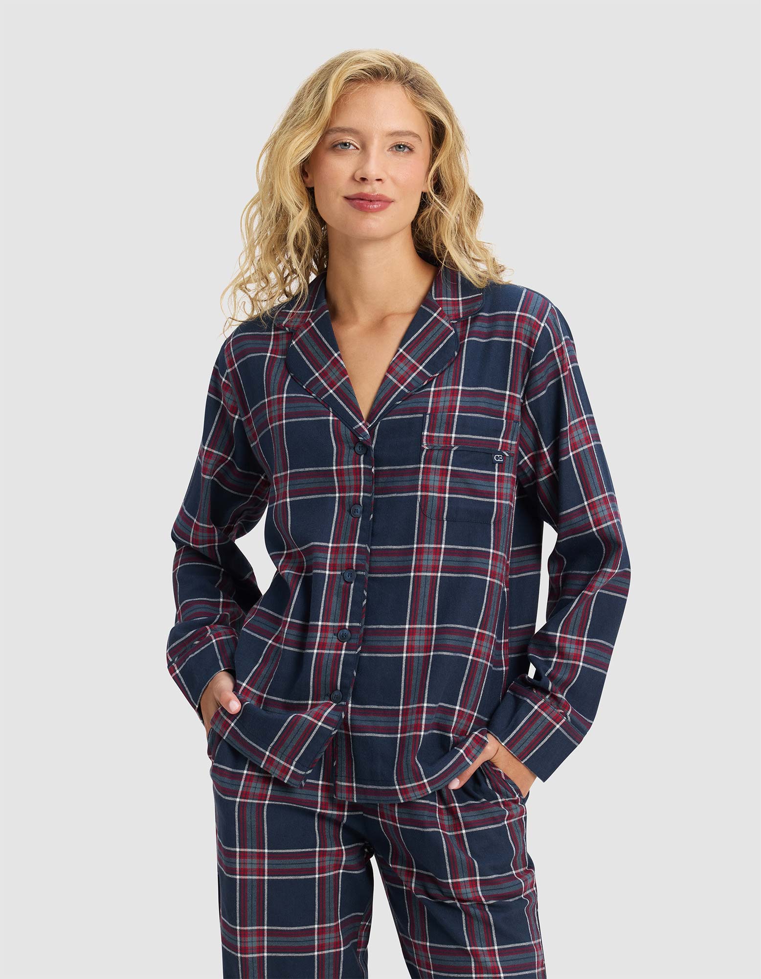 A woman with wavy blonde hair models the Cozy Earth Women's Farmhouse Long Sleeve Pajama Top in a navy, red, and white plaid pattern, standing with hands in her pockets and smiling against a plain light gray background.|Color:Eclipse Plaid