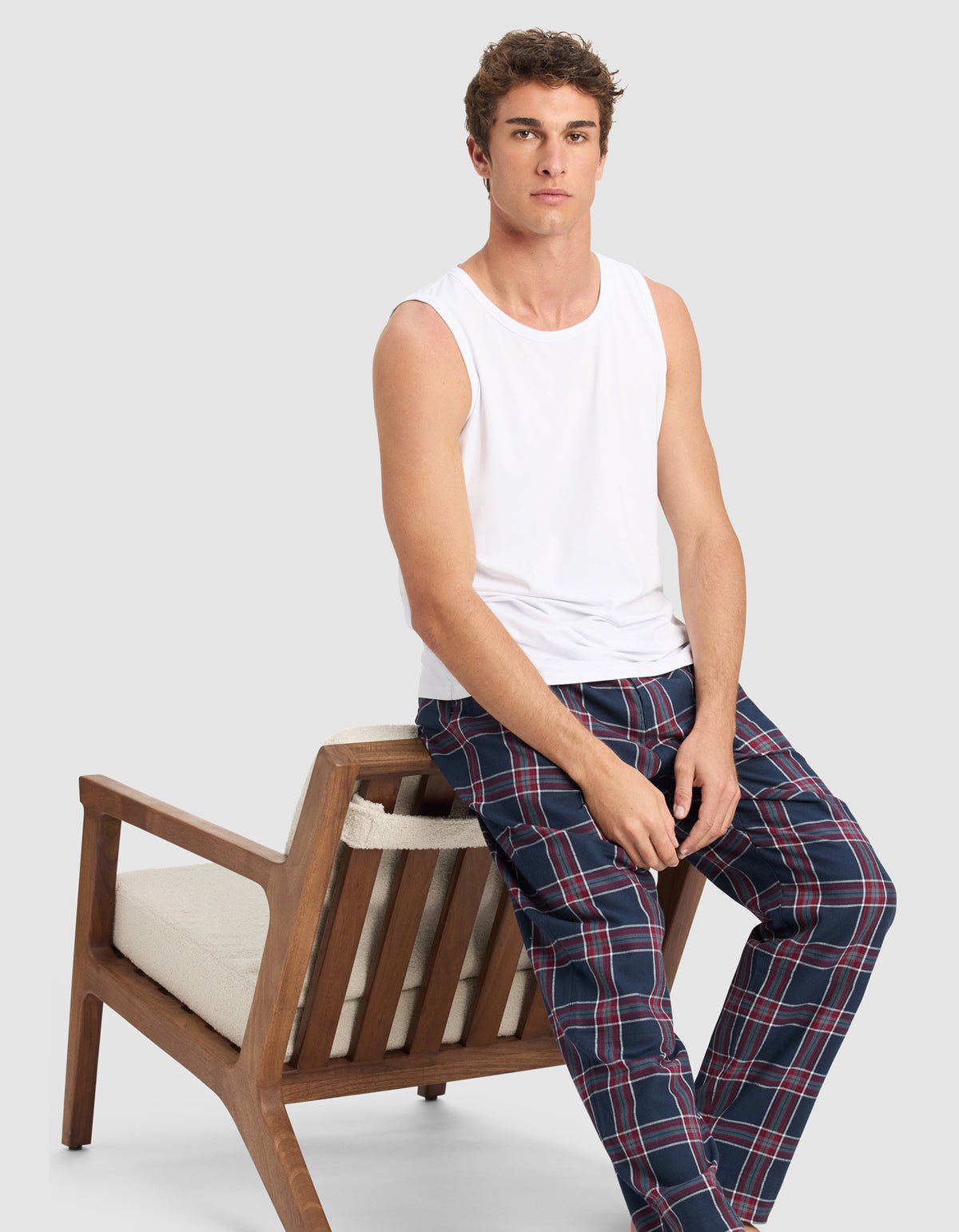 A young man in a white sleeveless shirt and Cozy Earth Men's Farmhouse Pajama Pant sits on the armrest of a wooden chair with a neutral expression against a light gray background.