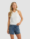 A woman with long blonde hair wears the Cozy Earth Women's Fine Ribbed Tank in white and blue denim shorts, standing against a light gray background and looking slightly to her right.|Color:Vapor