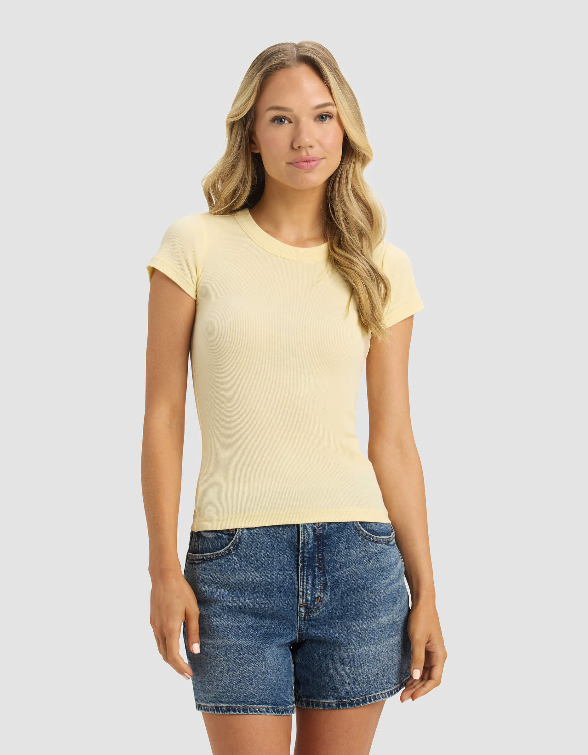 A woman with long blonde hair is wearing the Cozy Earth Women's Fine Ribbed Tee in light yellow and denim shorts, standing against a plain light grey background.|Color:Meringue