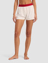 A person stands barefoot against a plain light background, wearing Cozy Earth Women's First Layer Pajama Boxer Shorts in white with small red polka dots and a red waistband. Their upper body is not shown.|Color:Cranberry Sea Salt Mini Heart
