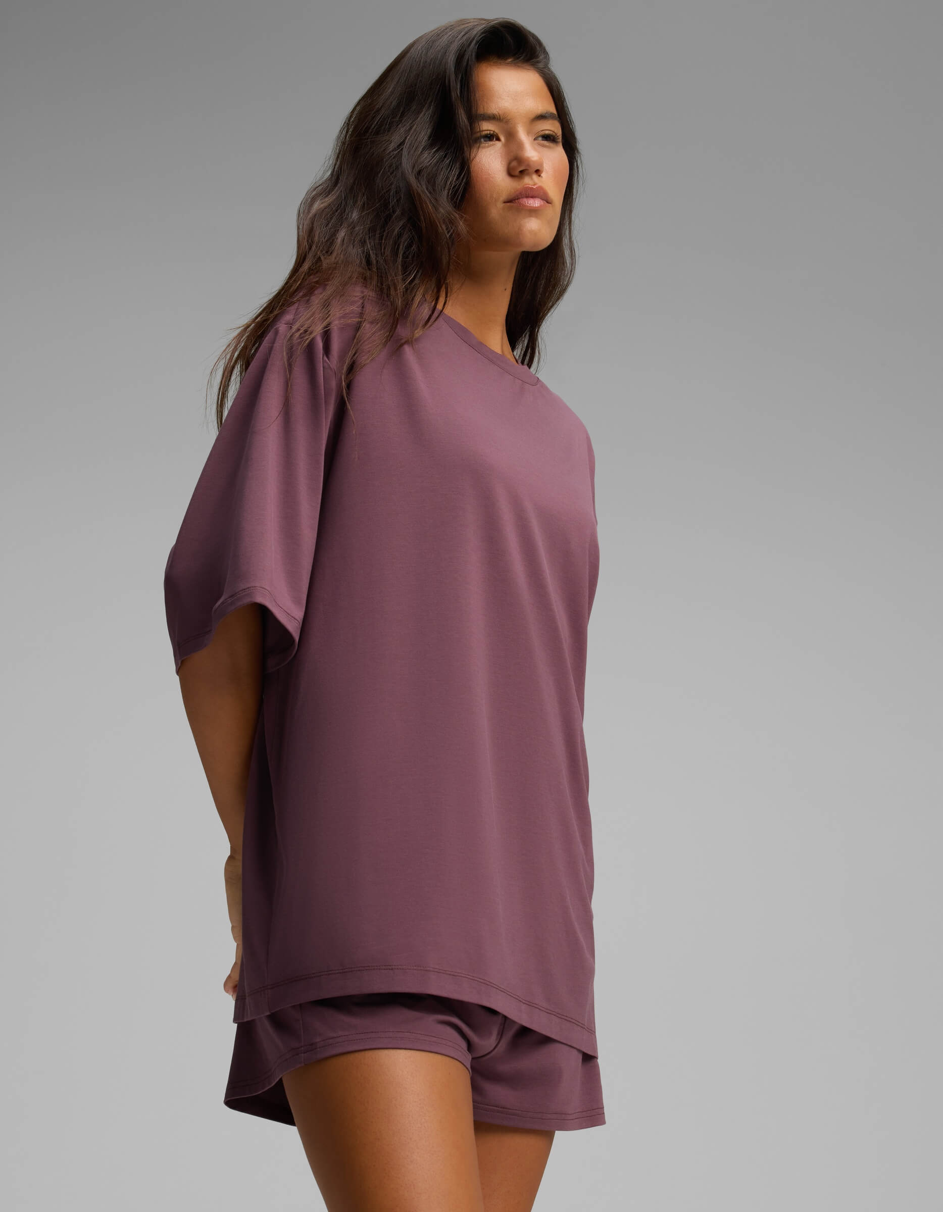 A woman with long dark hair is wearing the Cozy Earth Women’s First Layer Boyfriend Sleep Tee in purple, paired with matching shorts. She stands against a plain gray background, looking slightly to the side.|Color:Fig