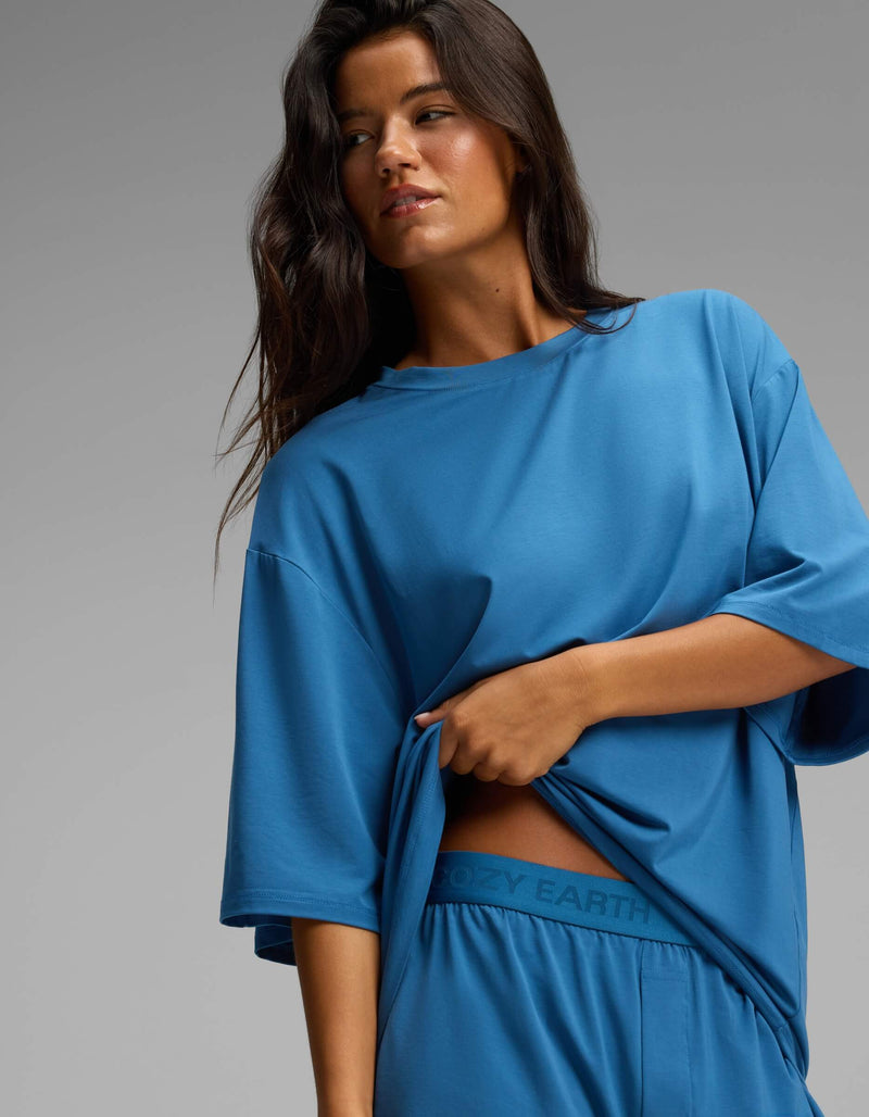 A woman with long dark hair gazes to the side against a plain gray background while lifting the hem of her loose blue Cozy Earth Women’s First Layer Boyfriend Sleep Tee to reveal matching blue Boxer Shorts featuring the “COZY EARTH” logo on the waistband.