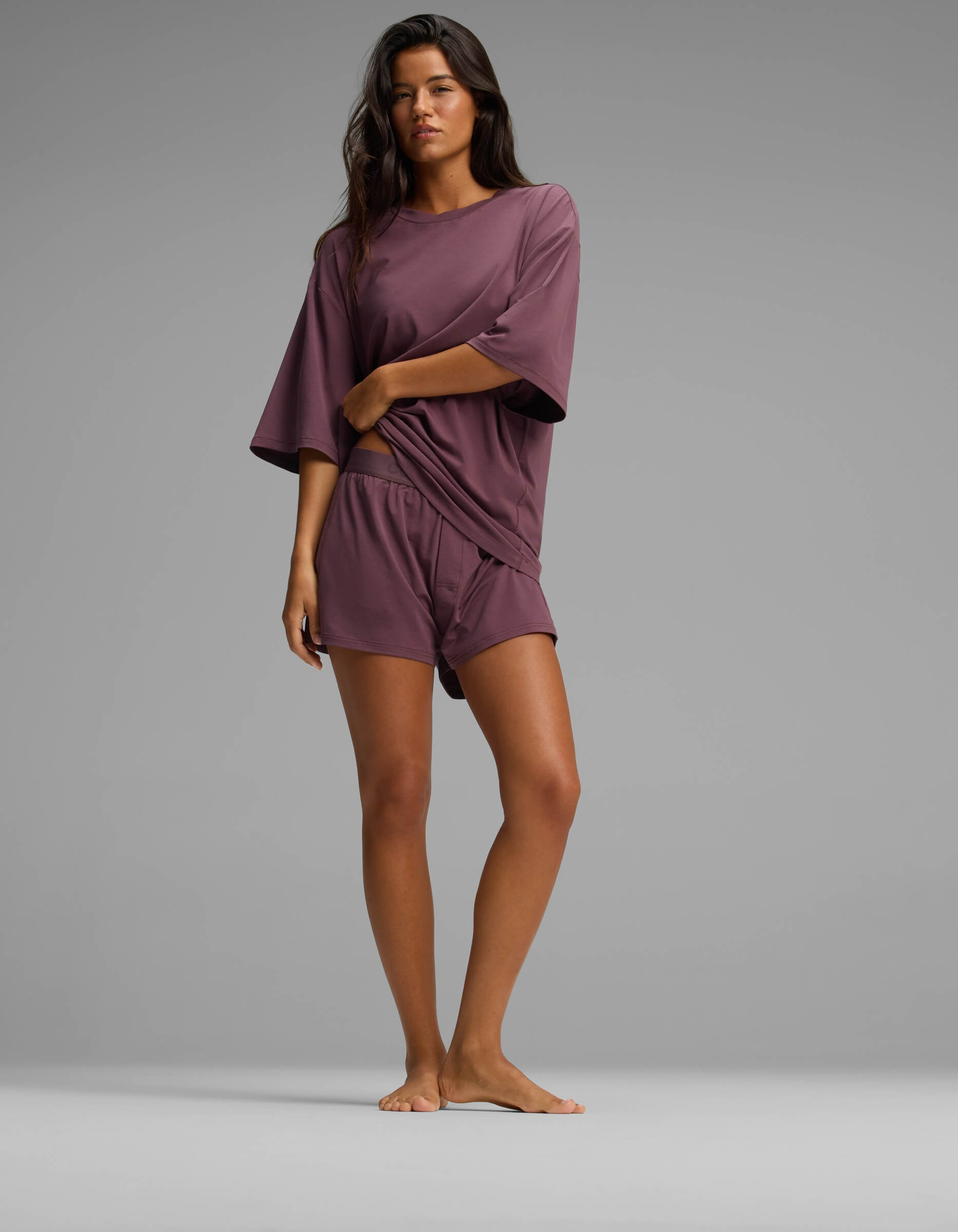 A woman stands barefoot on a plain background, wearing the Cozy Earth Women’s First Layer Boyfriend Sleep Tee in mauve with matching shorts, holding the hem of her shirt with one hand.