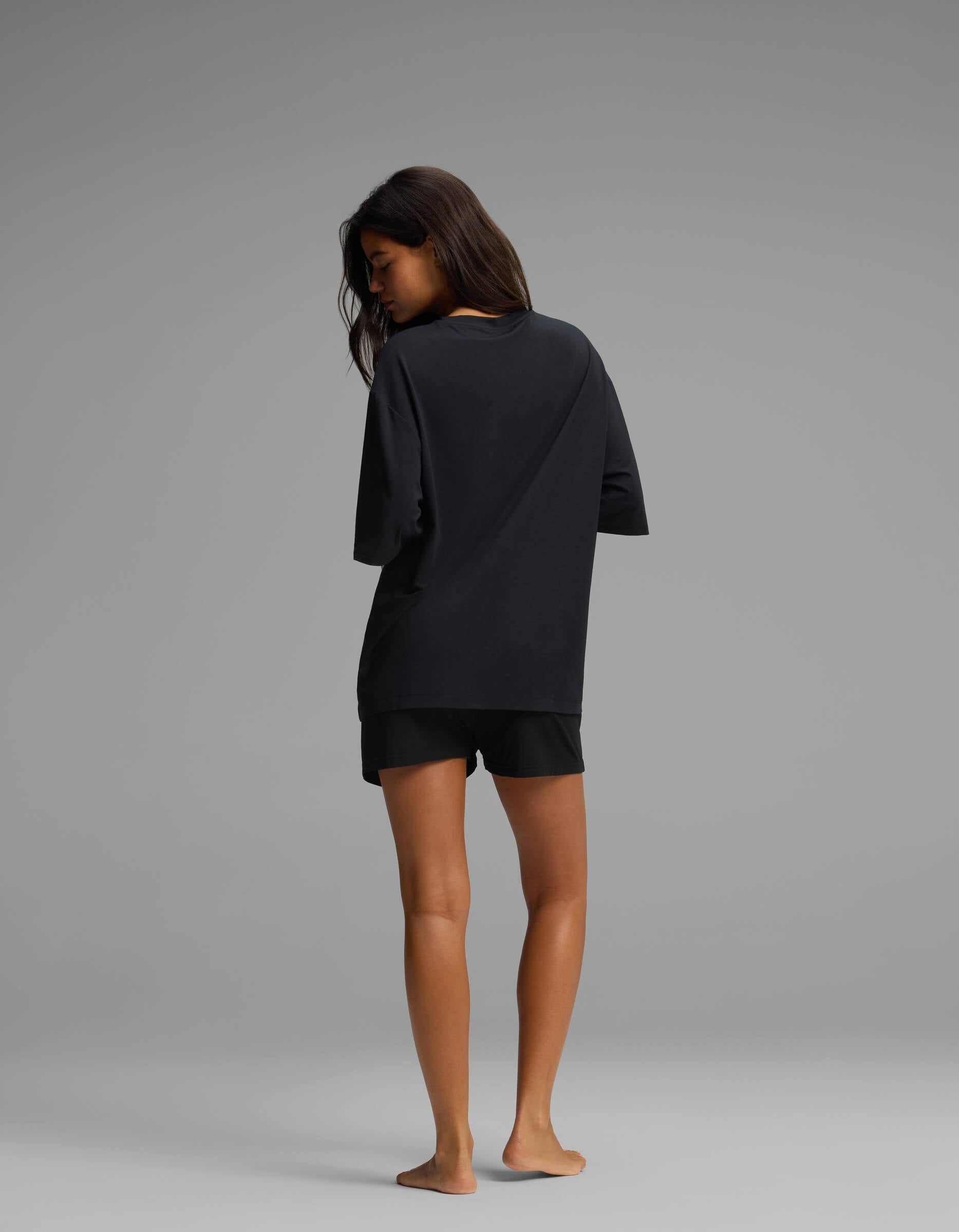 A person stands barefoot on a gray floor, facing away from the camera, wearing the Cozy Earth Women’s First Layer Boyfriend Sleep Tee & Boxer Short Set in black against a plain gray background.
