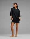 A woman stands barefoot against a gray background, wearing the Cozy Earth Women’s First Layer Boyfriend Sleep Tee & Boxer Short Set in a loose-fitting black fabric with small white dots.|Color:Mini Ghost