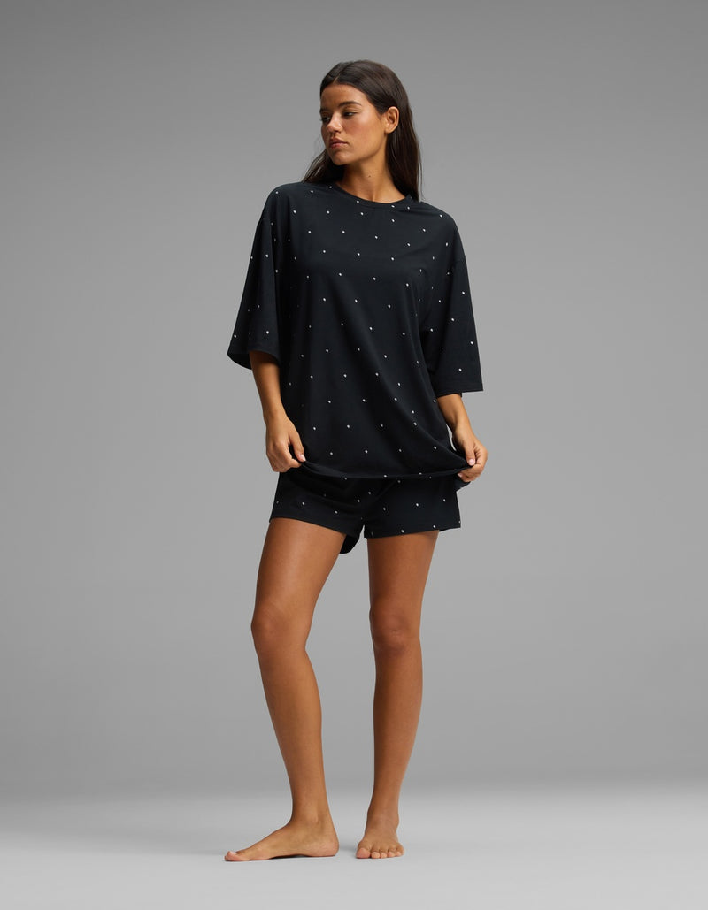 A woman stands barefoot against a gray background, wearing the Cozy Earth Women’s First Layer Boyfriend Sleep Tee & Boxer Short Set in a loose-fitting black fabric with small white dots.