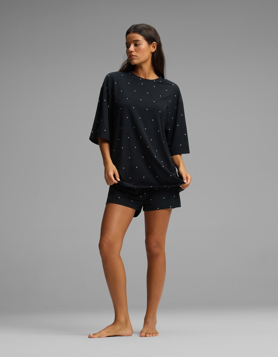 A woman stands barefoot against a gray background, wearing the Cozy Earth Women’s First Layer Boyfriend Sleep Tee & Boxer Short Set in a loose-fitting black fabric with small white dots.|Color:Mini Ghost