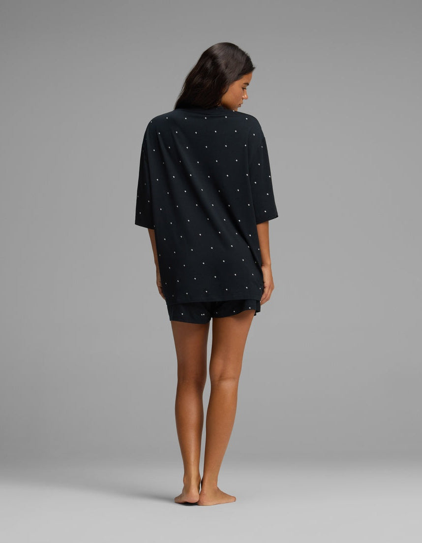 A woman stands barefoot on a gray background, facing away from the camera, wearing the Cozy Earth Women’s First Layer Boyfriend Sleep Tee & Boxer Short Set in a dark color with small white dots.