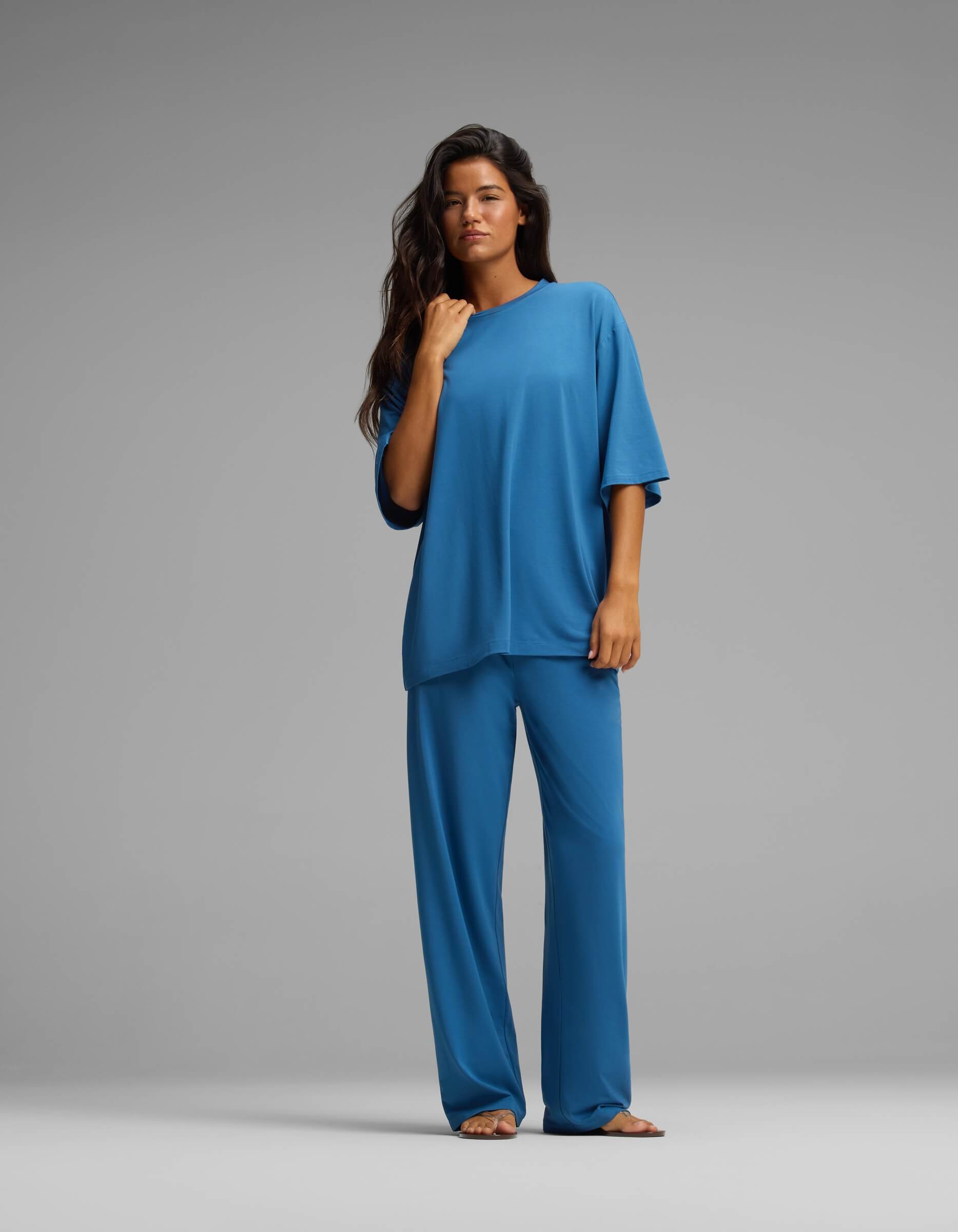 A long-haired woman stands against a grey background, wearing the Cozy Earth Women’s First Layer Boyfriend Sleep Tee & Pajama Pant Set—a loose-fitting, short-sleeved blue top paired with matching wide-leg blue pajama pants. She looks directly at the camera, one hand resting near her collarbone.|Color:Anchor