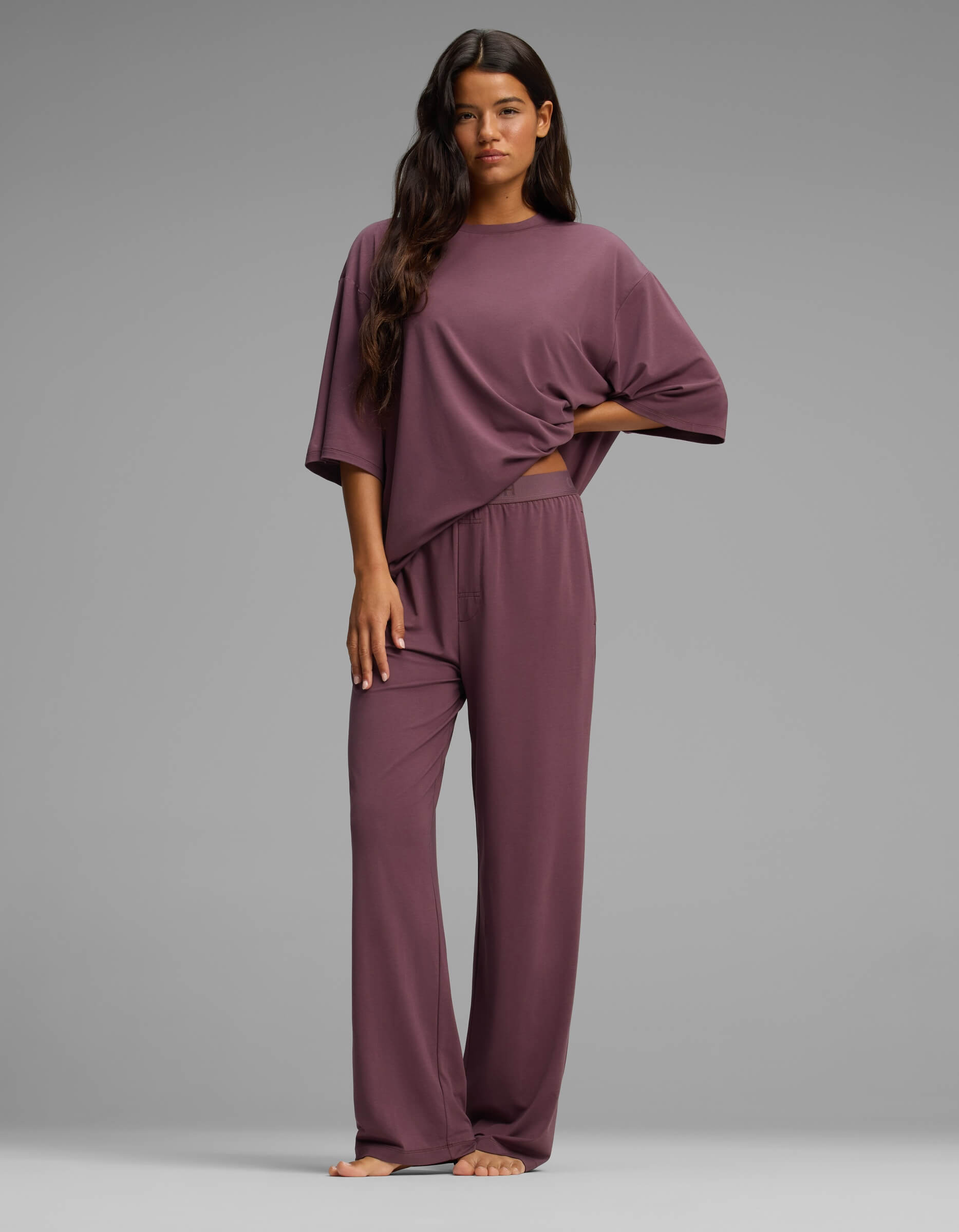 A woman stands barefoot against a gray background, wearing Cozy Earth’s Women’s First Layer Boyfriend Sleep Tee & Pajama Pant Set—a loose, mauve loungewear ensemble featuring a short-sleeve top and relaxed pants.|Color:Fig