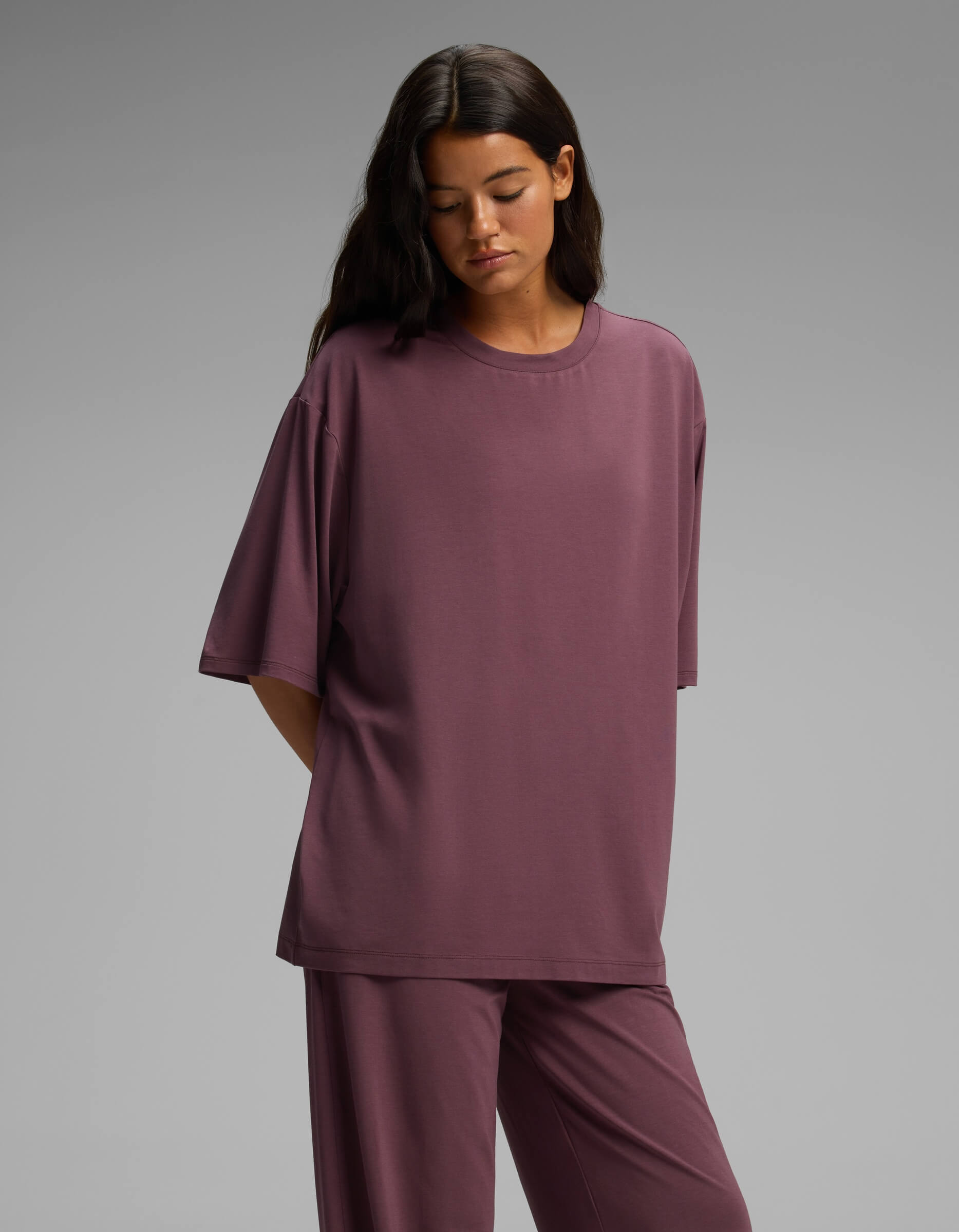 A woman with long dark hair stands against a plain gray background, her hands behind her back, wearing the Cozy Earth Women’s First Layer Boyfriend Sleep Tee & Pajama Pant Set in a loose mauve fit.
