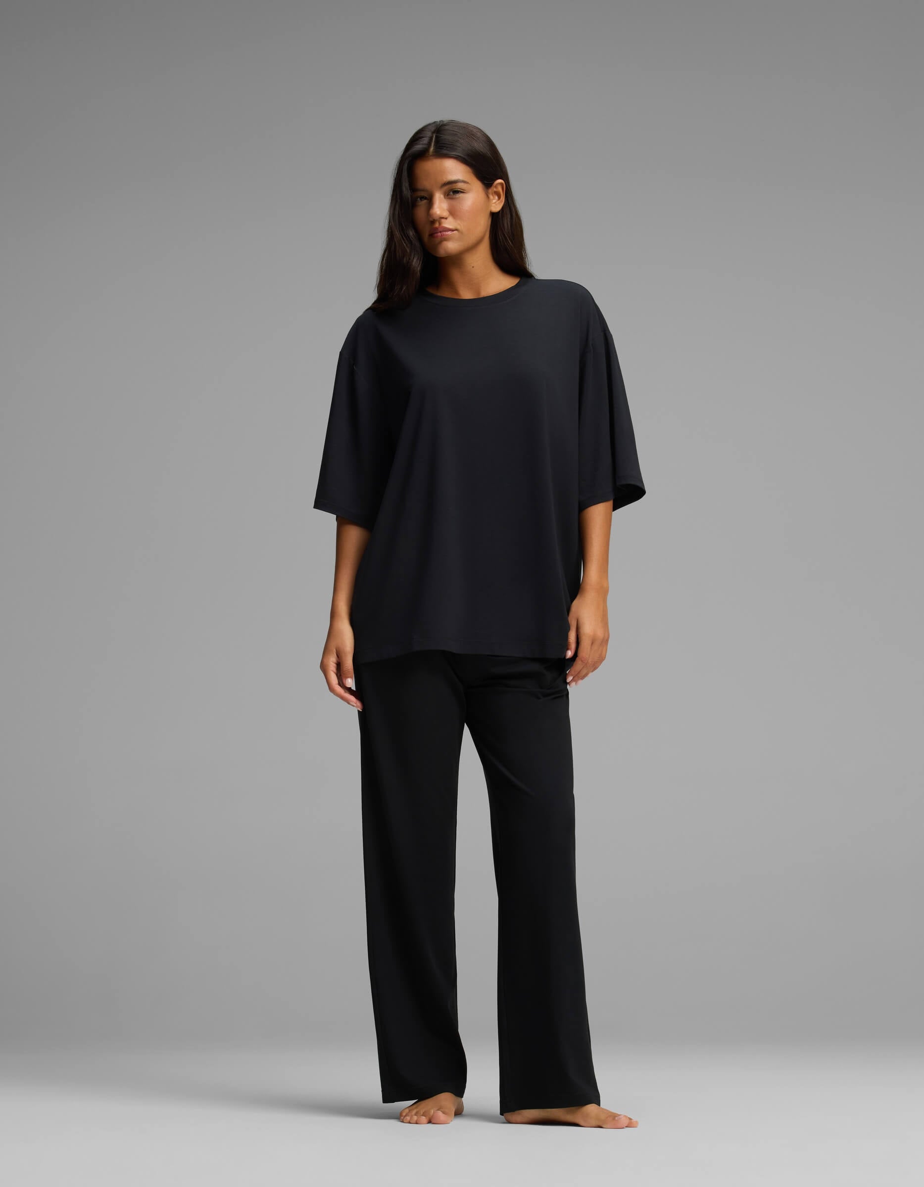 A woman stands barefoot against a gray background, wearing the Cozy Earth Women’s First Layer Boyfriend Sleep Tee & Pajama Pant Set in black, with her hair down and her arms relaxed by her sides.