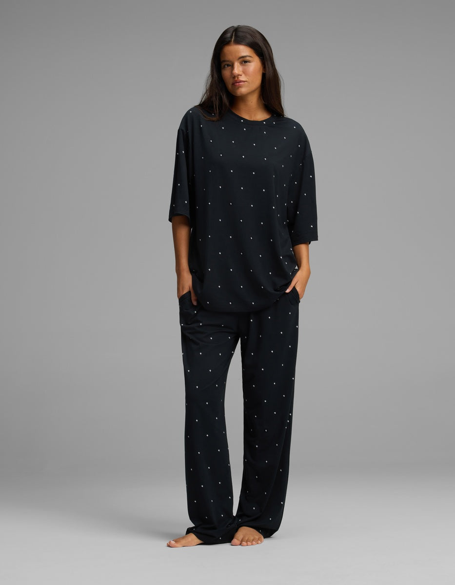 A woman stands barefoot in a relaxed pose, wearing the Cozy Earth Women’s First Layer Boyfriend Sleep Tee & Pajama Pant Set—a loose-fitting black pajama ensemble with small white polka dots—against a plain gray background.