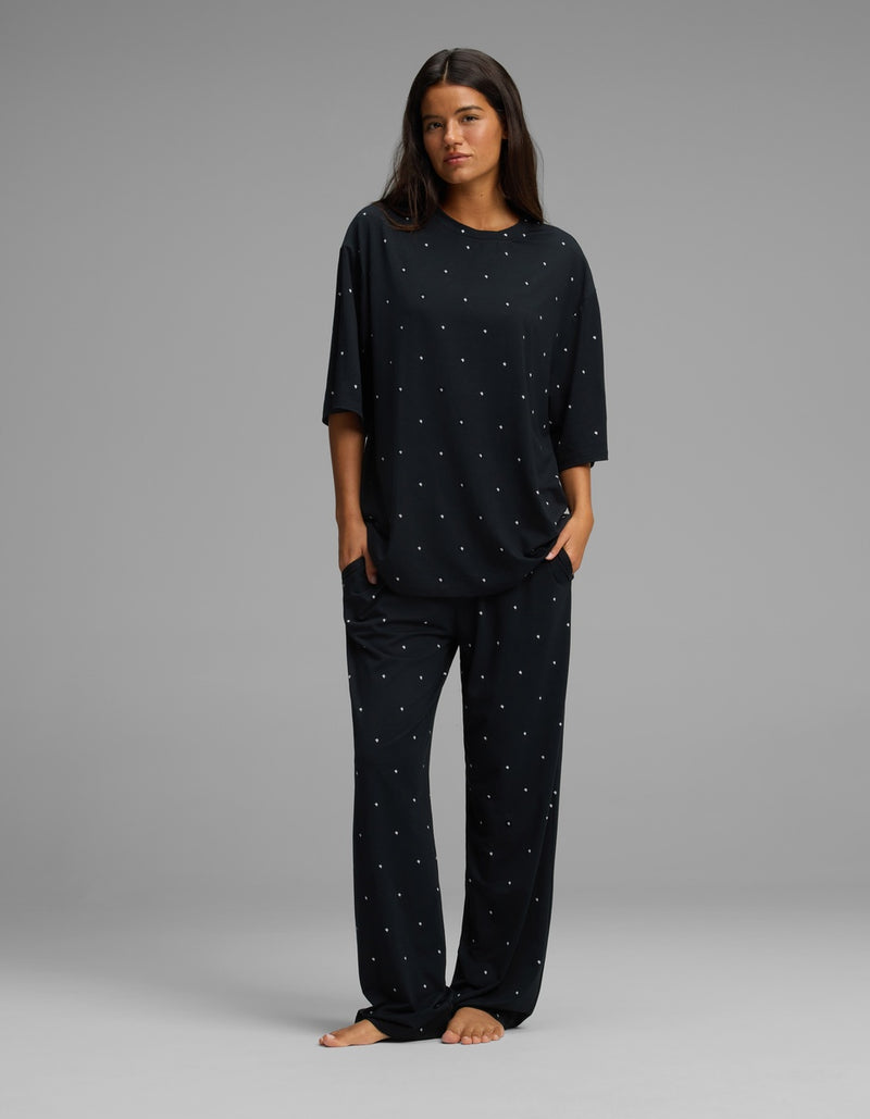 A woman stands barefoot in a relaxed pose, wearing the Cozy Earth Women’s First Layer Boyfriend Sleep Tee & Pajama Pant Set—a loose-fitting black pajama ensemble with small white polka dots—against a plain gray background.