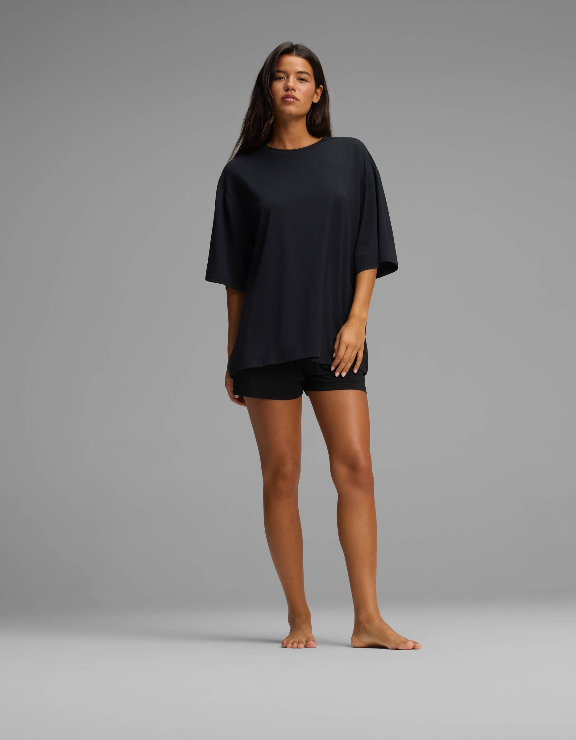 A woman stands barefoot against a plain gray background, wearing the Cozy Earth Women’s First Layer Boyfriend Sleep Tee in black and matching black shorts, with her right hand resting on her thigh and her left arm relaxed by her side.