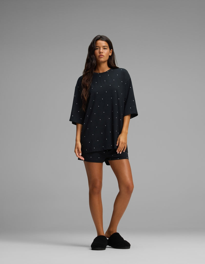 A woman with long brown hair and a neutral expression stands against a gray background wearing the Cozy Earth Women’s First Layer Boyfriend Sleep Tee in black, paired with matching black shorts and black slippers.