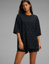 A woman stands against a plain gray background, wearing the Cozy Earth Women’s First Layer Boyfriend Sleep Tee in black with small white polka dots, paired with matching shorts.|Color:Mini Ghost