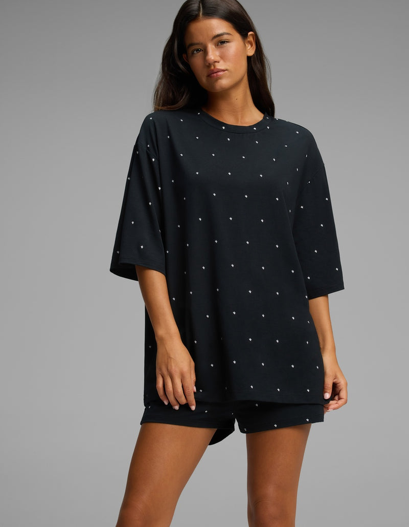 A woman stands against a plain gray background, wearing the Cozy Earth Women’s First Layer Boyfriend Sleep Tee in black with small white polka dots, paired with matching shorts.
