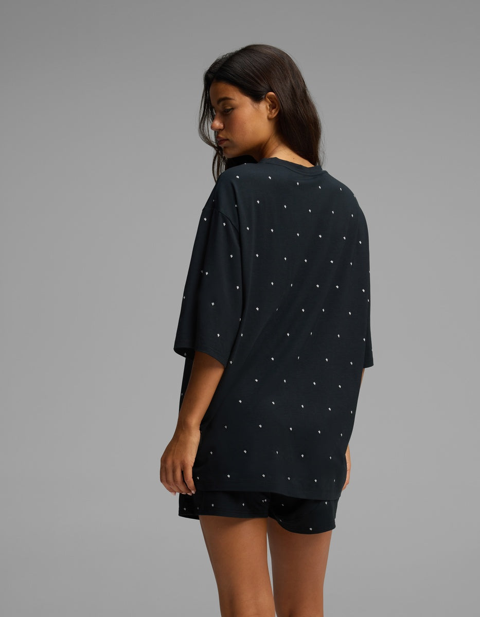 A woman with long dark hair faces away, wearing the Cozy Earth Women’s First Layer Boyfriend Sleep Tee in black, paired with matching shorts featuring small white dots. The background is plain gray.|Color:Mini Ghost
