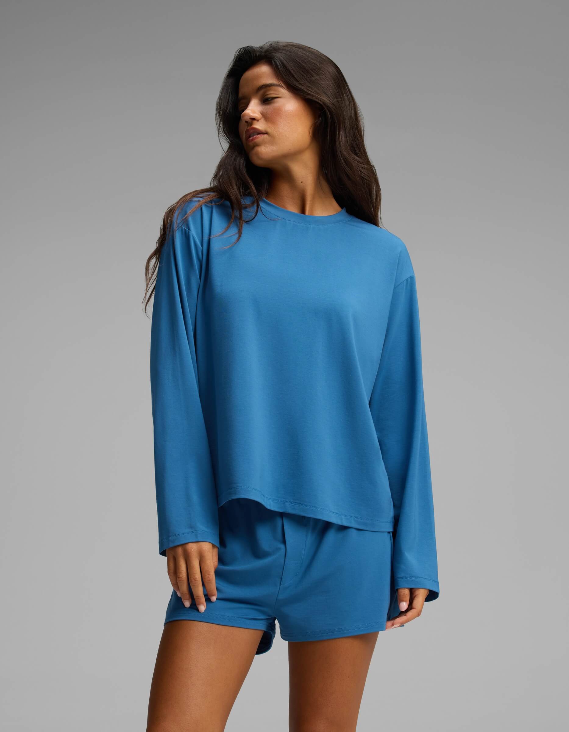 A woman wearing the Cozy Earth Women’s First Layer Long Sleeve Sleep Tee in blue and coordinating shorts stands against a plain gray background, looking to the side with her hand relaxed by her leg.