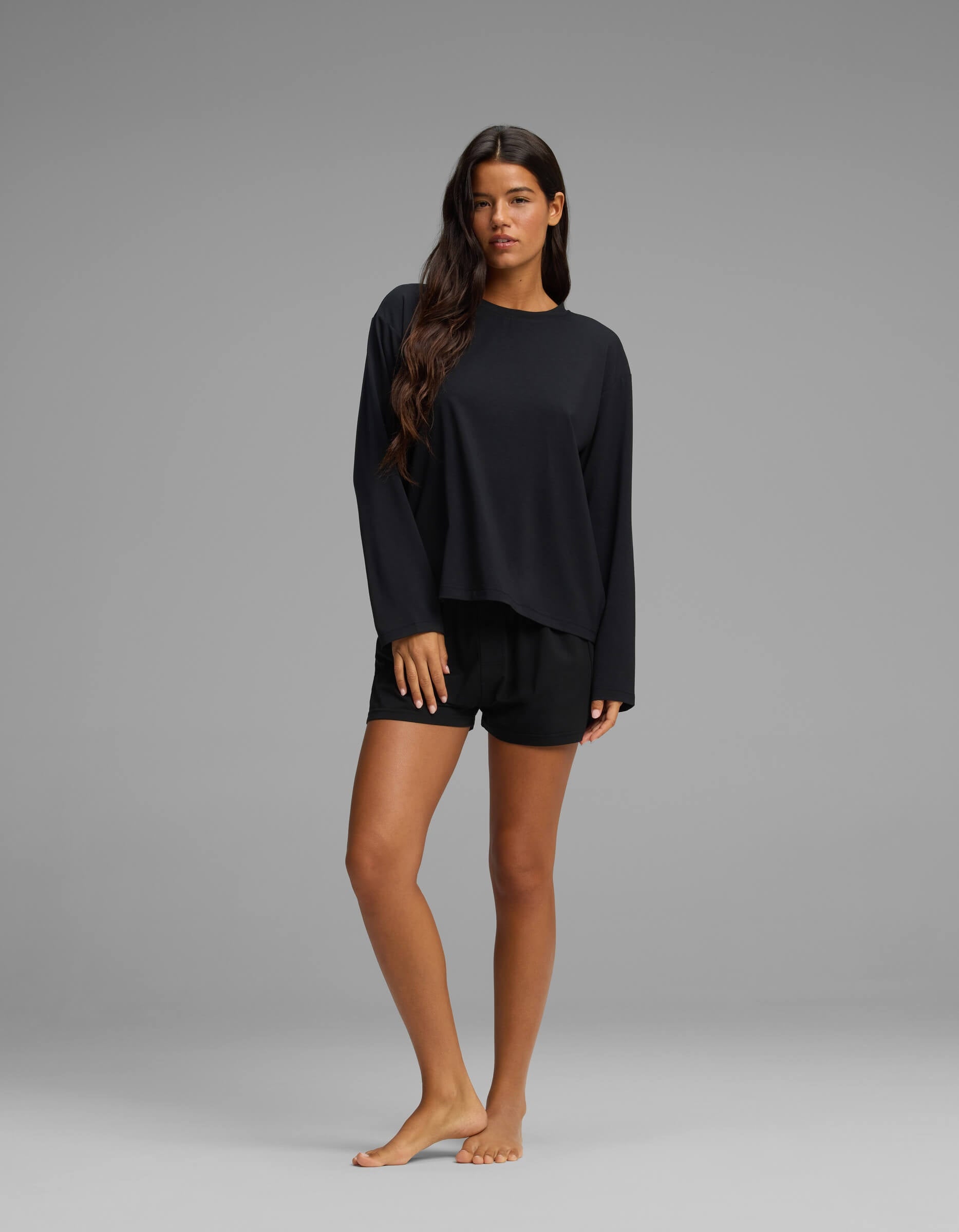 A woman with long brown hair stands barefoot on a grey background, modeling the Cozy Earth Women’s First Layer Long Sleeve Sleep Tee & Boxer Short Set in black, looking at the camera with a neutral expression.