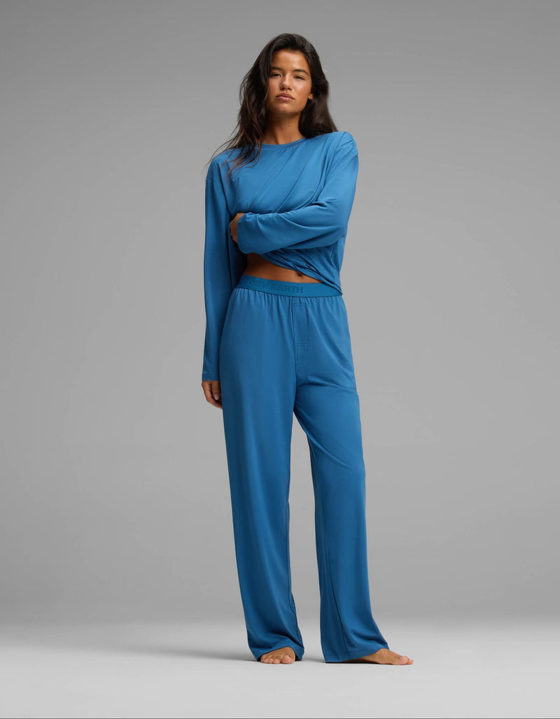 A person stands barefoot against a plain gray background, wearing the Cozy Earth Women’s First Layer Long Sleeve Sleep Tee & Pajama Pant Set in blue. They have long dark hair and a neutral expression, with one arm crossed over their body.