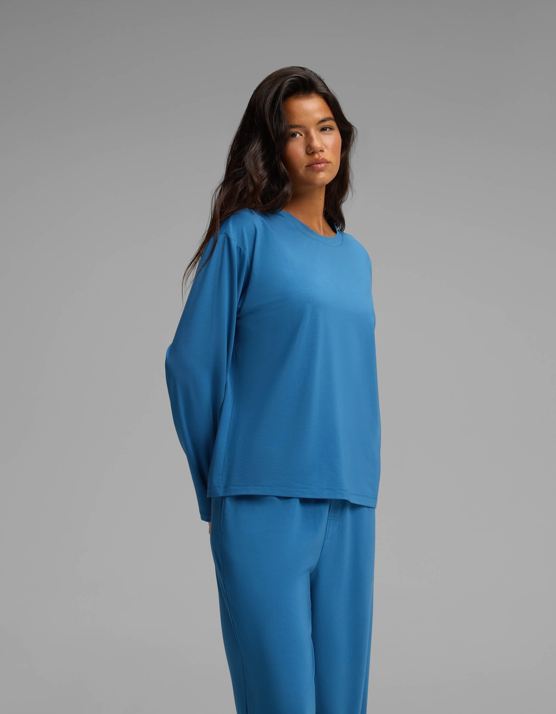 A woman with long dark hair wearing a loose blue long-sleeve top and matching blue pants stands against a plain gray background.