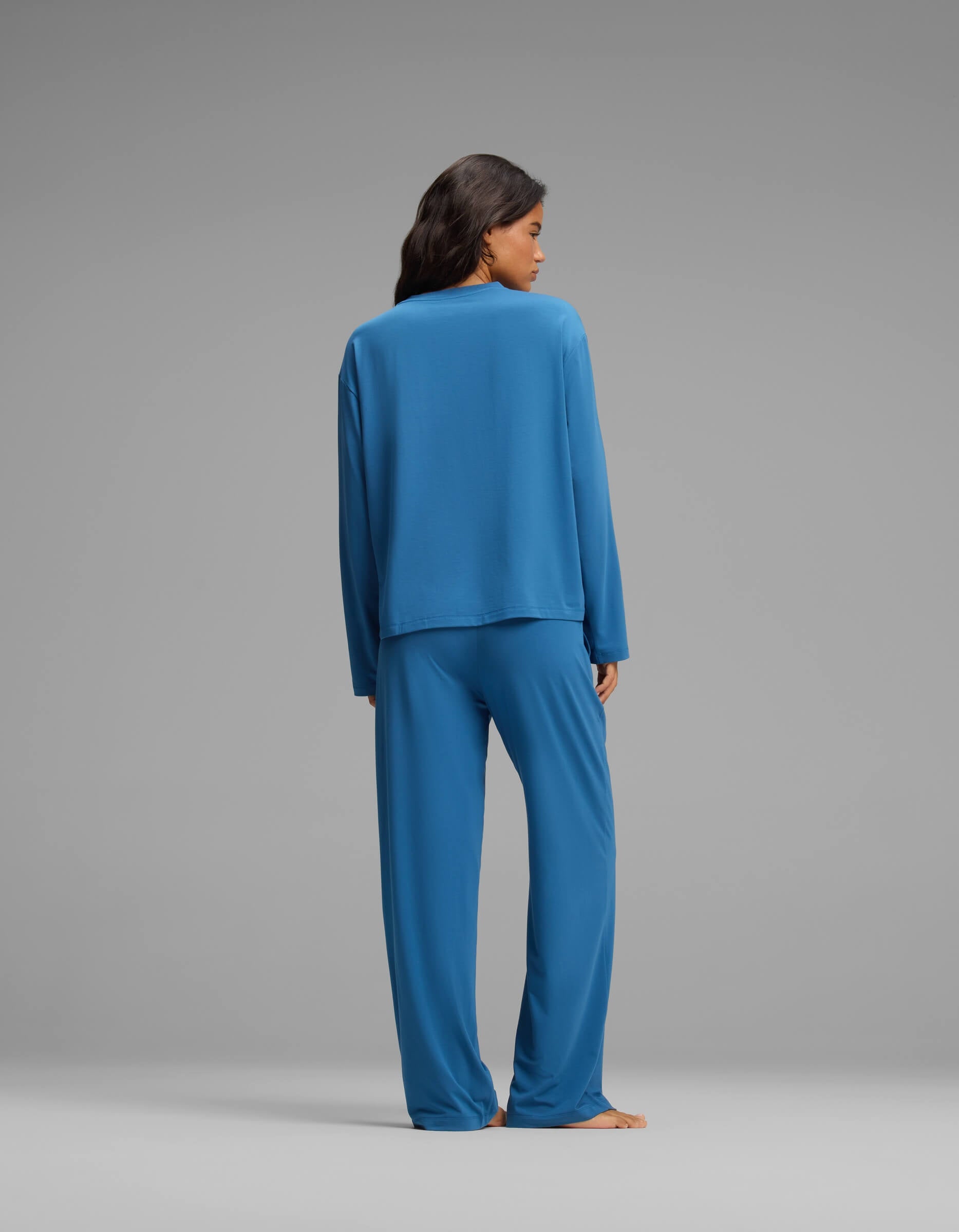 A woman with dark hair stands with her back to the camera against a plain gray background, wearing the Cozy Earth Women’s First Layer Long Sleeve Sleep Tee & Pajama Pant Set in blue.