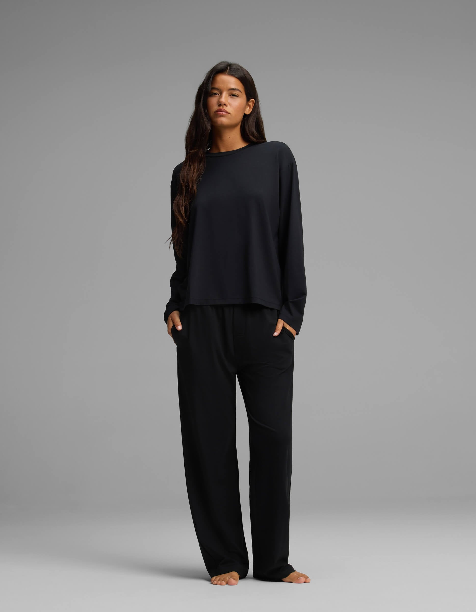 A woman with long brown hair stands facing forward against a plain gray background. She is wearing the Cozy Earth Women’s First Layer Long Sleeve Sleep Tee & Pajama Pant Set in black, with her hands tucked into the side pockets. She is barefoot.