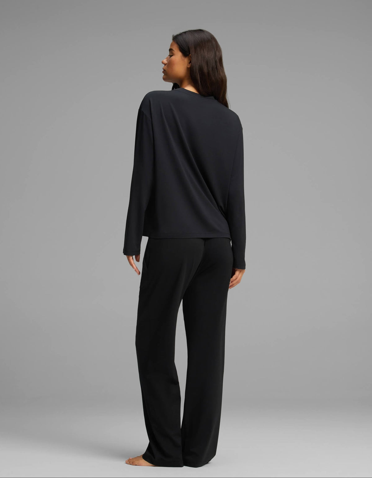 A woman with long brown hair, seen from behind, stands barefoot against a plain gray background wearing the Cozy Earth Women’s First Layer Long Sleeve Sleep Tee & Pajama Pant Set in black.|Color:Jet Black