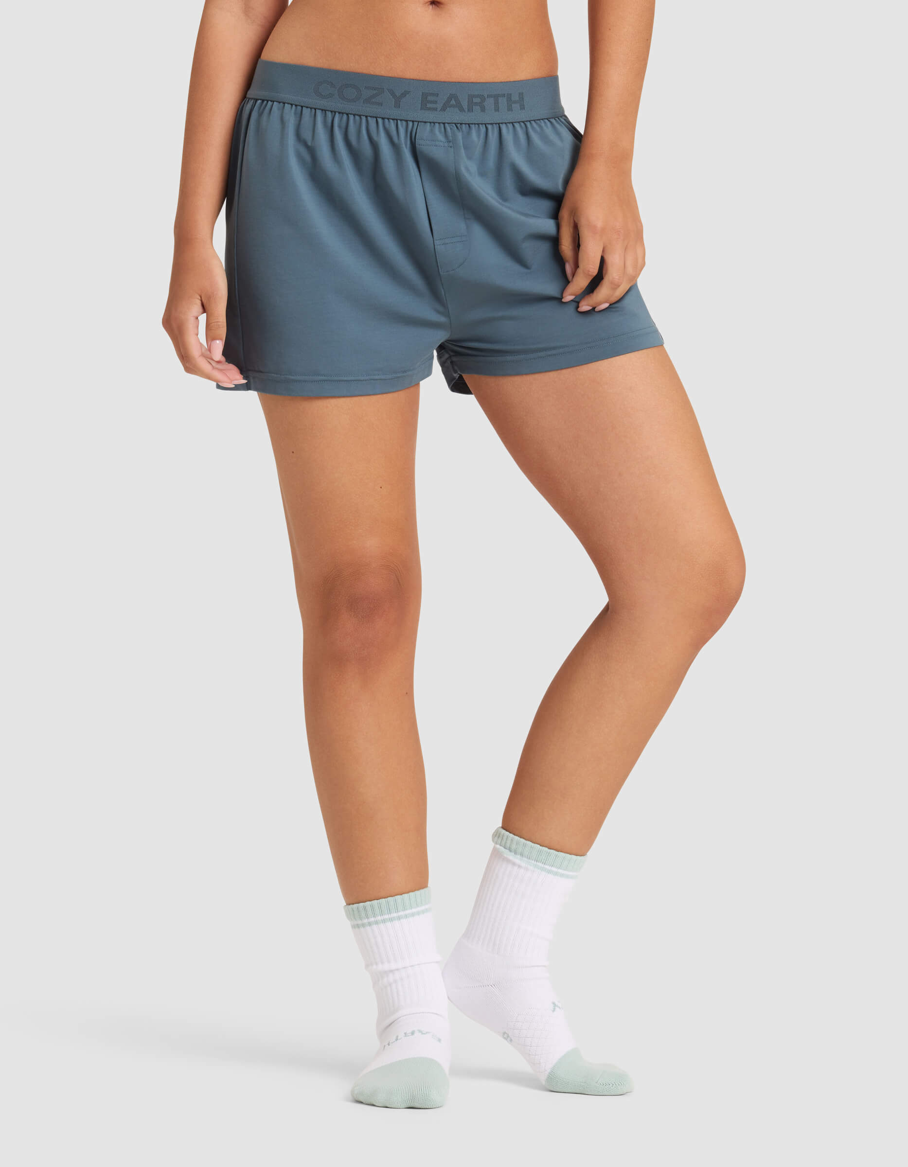 A person is shown from the waist down wearing Cozy Earth Women's First Layer Pajama Boxer Shorts in blue-gray, featuring the brand logo on the waistband, paired with white socks and a white top, set against a plain light background.|Color:Dawn