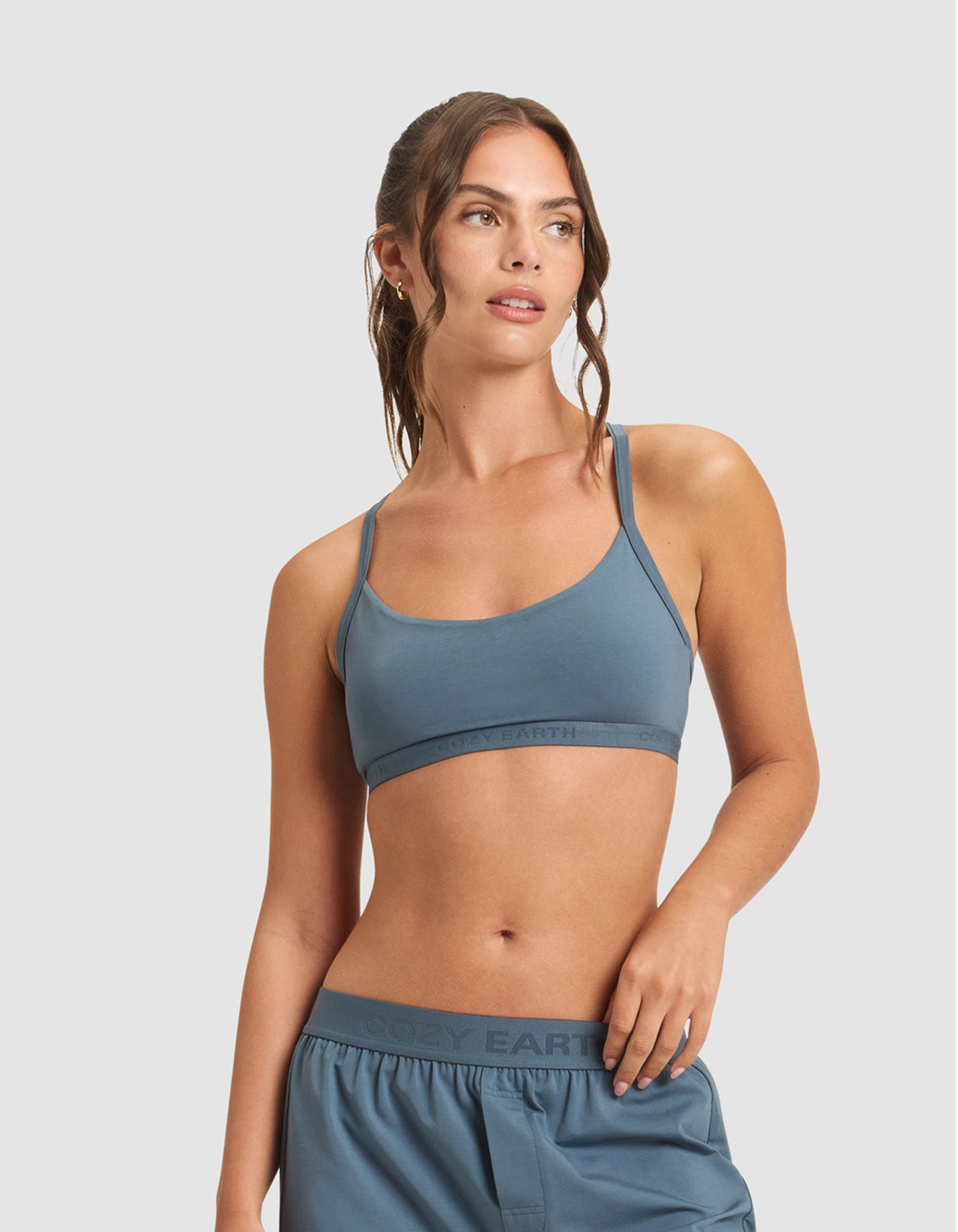 A woman with wavy brown hair in a ponytail wears the Cozy Earth Women's First Layer Pajama Soft Bra and matching shorts, looking to the side against a plain light gray background.|Color:Dawn