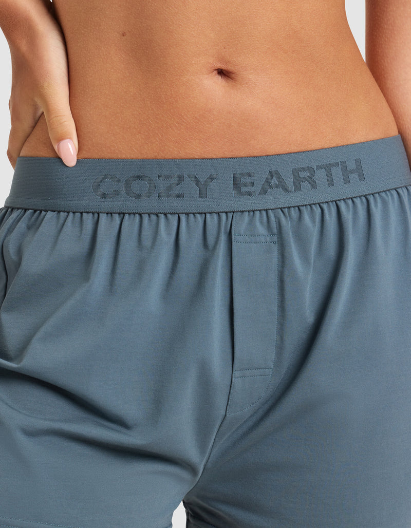 A close-up shows a person wearing Cozy Earth Women’s First Layer Pajama Boxer Shorts in blue-gray, with the branded waistband visible and their hand resting on their hip.