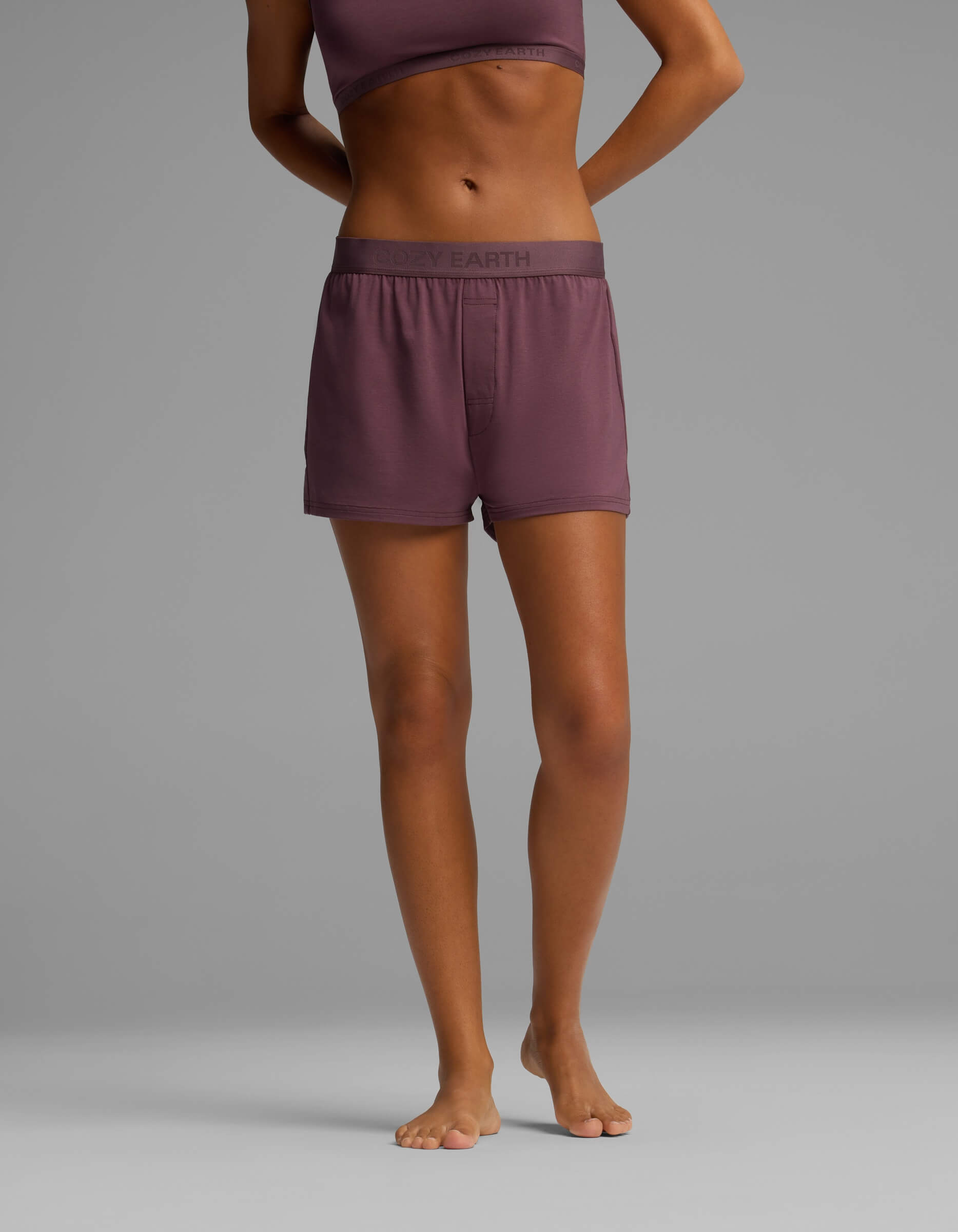 A person standing barefoot against a plain gray background, wearing Cozy Earth’s Women’s First Layer Pajama Boxer Short in mauve with a matching sports bra. Only the body from shoulders to feet is visible.
