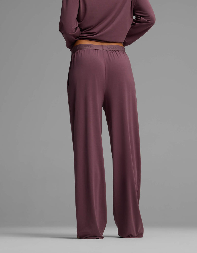 A woman is seen from the back wearing Cozy Earth’s Women’s First Layer Pajama Pant in a loose mauve fit, paired with a matching long-sleeve top, set against a plain gray background.
