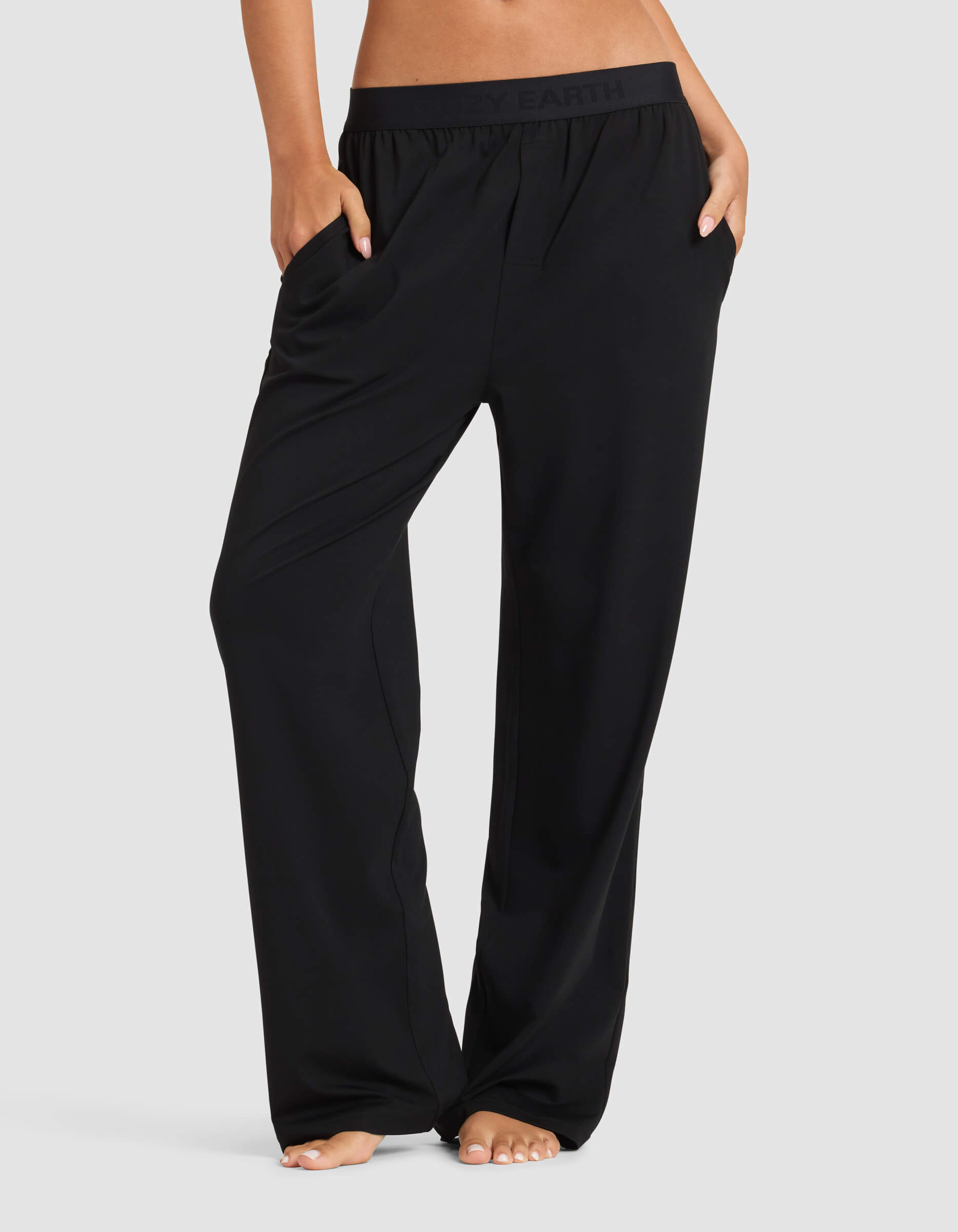 A person stands barefoot against a plain light background, wearing Cozy Earth’s Women's First Layer Pajama Pant in black, showcasing the relaxed fit and waistband with hands in pockets.|Color:Jet Black