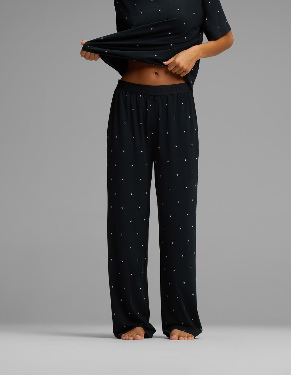 A person stands barefoot against a plain gray background, wearing the Cozy Earth Women’s First Layer Pajama Pant and a matching loose black top with small white polka dots, lifting the shirt slightly to reveal their midriff.|Color:Mini Ghost