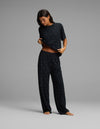 A woman stands barefoot on a gray background, wearing Cozy Earth’s Women’s First Layer Pajama Pant in a black fabric with small white dots. She slightly lifts her shirt to show her stomach while looking down.|Color:Mini Ghost